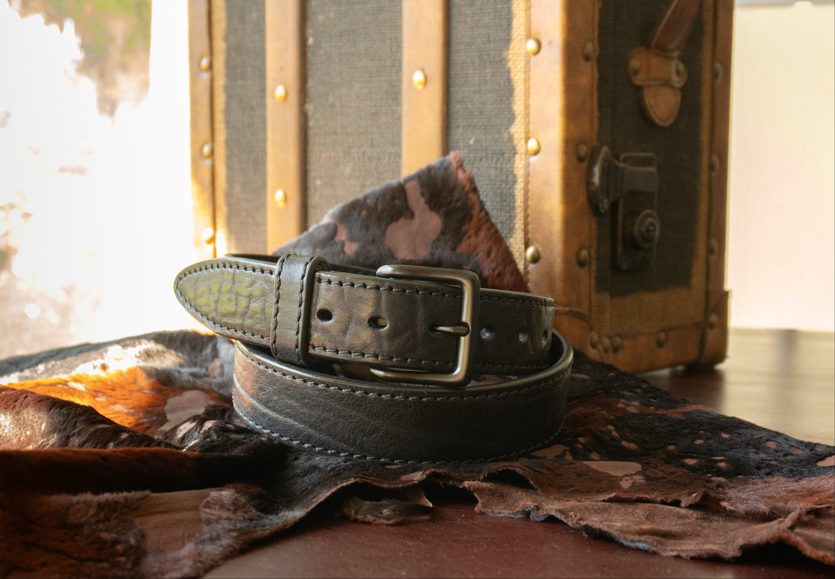 Black leather belt with a camouflage pattern on a textured surface with a vintage suitcase in the background.